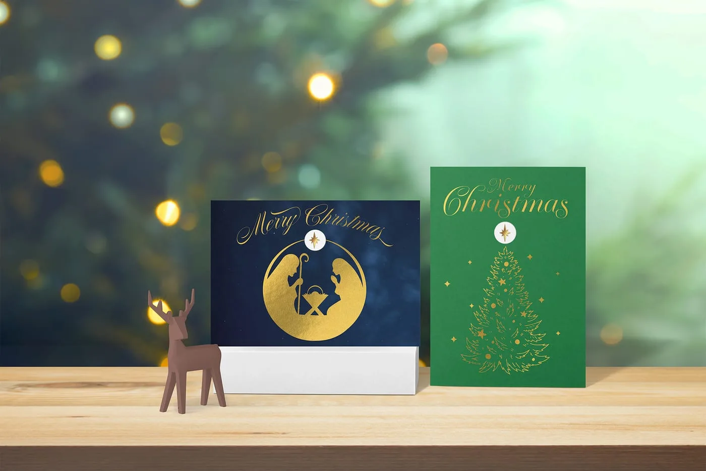 Lifestyle shot of the navy Nativity and green tree cards on a wooden surface before a lit Christmas tree